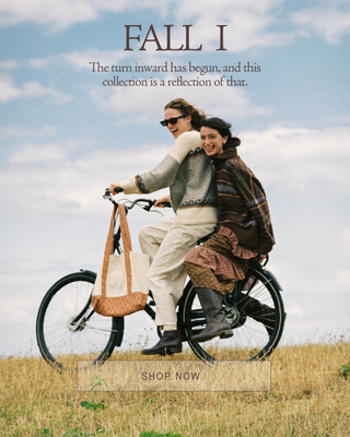 A lifestyle promotional image featuring two women riding a black bicycle outdoors on a grassy field under a cloudy sky. One woman wears a light gray knit cardigan and cream pants, while the other wears a dark brown knit sweater and a brown backpack.