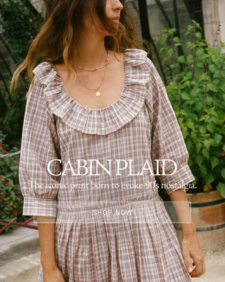 A woman stands in a garden wearing a light pink and white plaid dress, holding a matching plaid jacket. The background features a large historic stone building and manicured hedges, with the text 'CABINPLAID' and 'The iconic plaid: born to evoke 90s nostalgia.