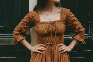 A woman wearing a rust-brown, long-sleeved midi dress with a smocked bodice, tie-front neckline, and ruffled cuffs, standing in front of a dark wooden door.