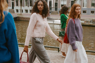 Two women walking along a canal-side walkway in Amsterdam, wearing cozy knit sweaters. One wears a pale pink button-front cardigan with gray trousers, and the other wears a lavender sweater with a white skirt.