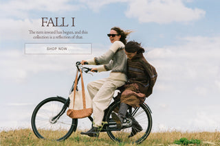 A lifestyle promotional image featuring two women riding a black bicycle outdoors on a grassy field under a cloudy sky. One woman wears a light gray knit cardigan and cream pants, while the other wears a dark brown knit sweater and a brown backpack.