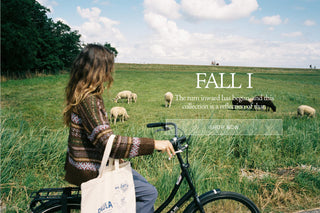 A lifestyle promotional image featuring a woman riding a black bicycle outdoors on a grassy field under a cloudy sky. woman wears a dark brown knit sweater and copy overlay says Fall 1 the turn inward has begun and this collection is a reflection of that. Shop Now