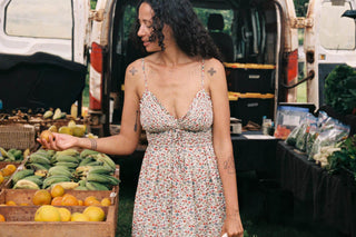 Woman in a floral dress standing at an outdoor farmers market with produce.