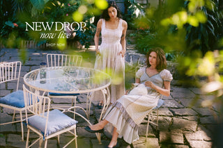 Two women in light dresses sitting at a glass table outdoors with 'New Drop Now Live and Shop Now' text overlay.