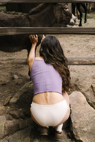 Woman in a purple top and white underwear leaning against a wooden fence with animals in the background.