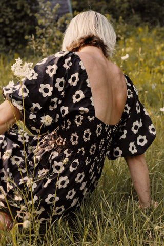 Person wearing a black dress with white floral pattern sitting in a field of grass and wildflowers.