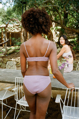 Two women in light purple lingerie standing outdoors near a wooden table with chairs.