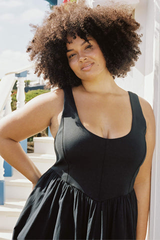 A woman with curly hair wearing a black sleeveless maxi dress with pockets.