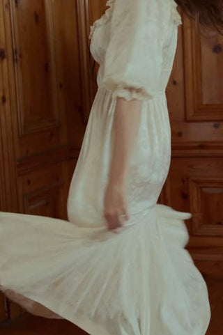 A woman wearing a flowing cream-colored dress with ruffled short sleeves, captured mid-motion as she twirls in front of a warm wooden paneled wall.