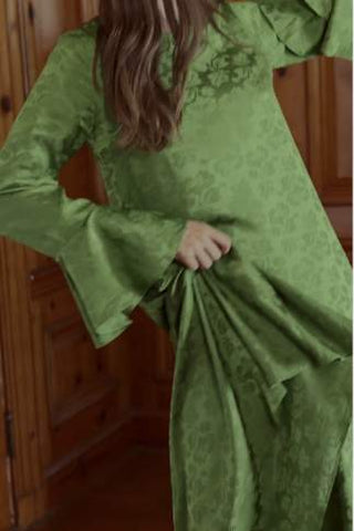 A woman wearing a green long-sleeved tunic dress with a subtle jacquard pattern, shown from the back in a warm indoor setting with wooden doors.