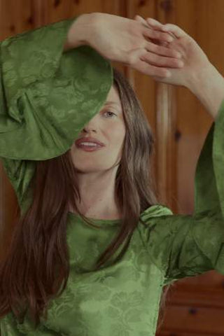 A woman with long brown hair wearing a green patterned long-sleeve top, posing with her arms raised in front of a warm wood-paneled background.