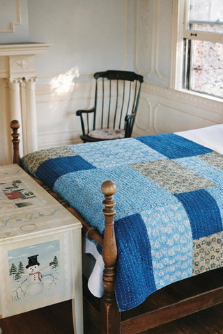 A quilt with various blue squares lays on a bed in a room with a rocking chair, window, and mantel piece