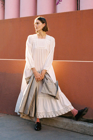 Woman in a long white dress holding a gray bag against a pink wall.

