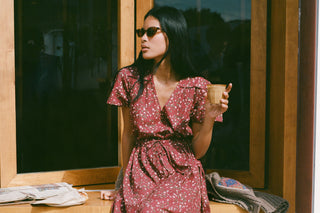 Woman in a pink dress holding a cup, sitting by a window.