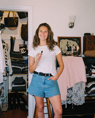 A woman stands on a wooden stool in a closet-filled room, wearing a white t-shirt and blue denim shorts. She is holding a small black object in her right hand.