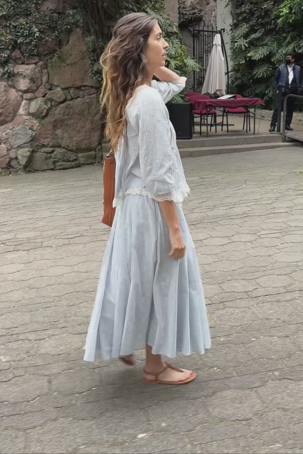 Woman in a light blue outfit walking on a street with plants behind her