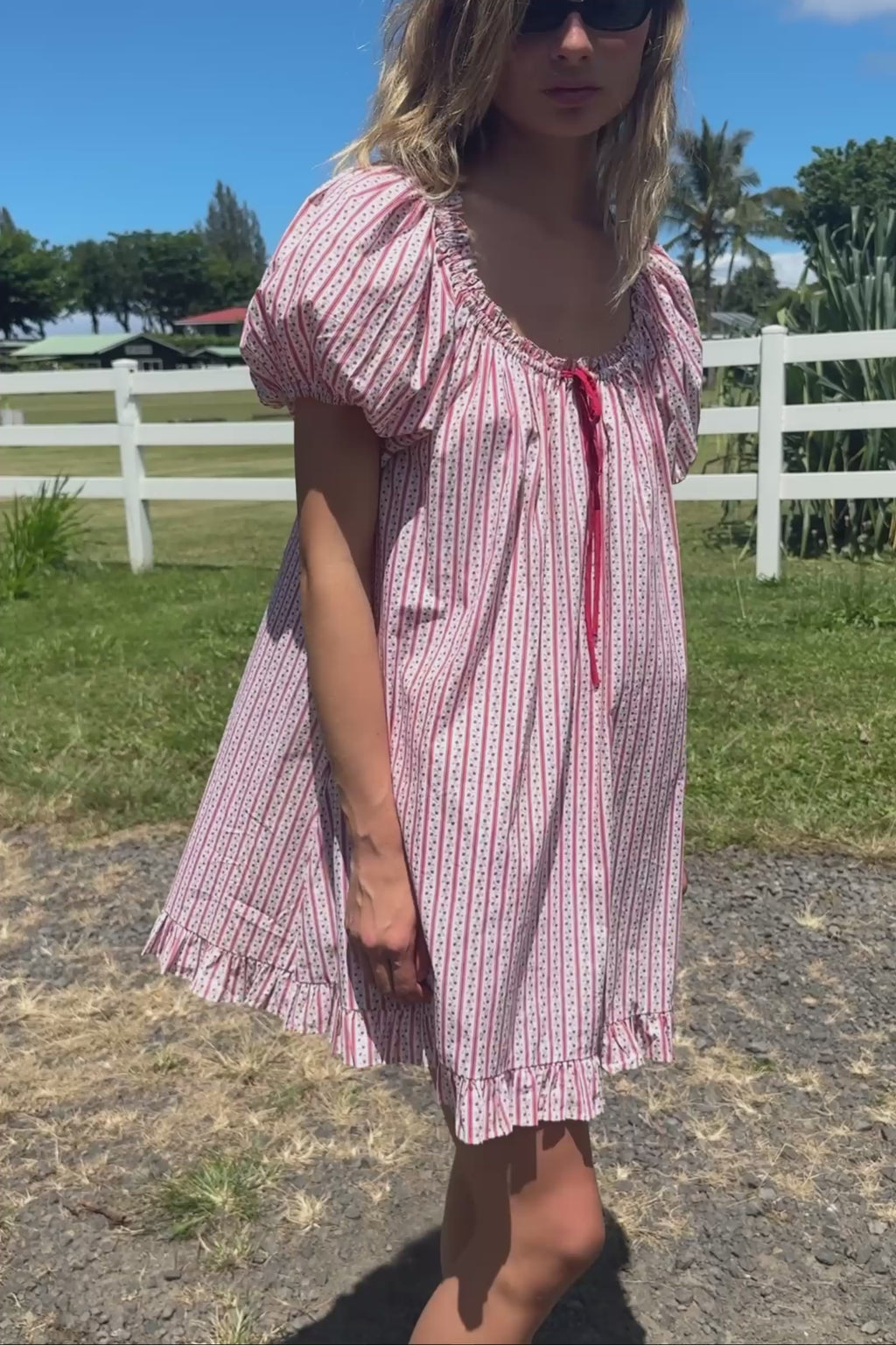 A video of a woman in a red and white striped pattern dress standing in grass with a white fence in the background.