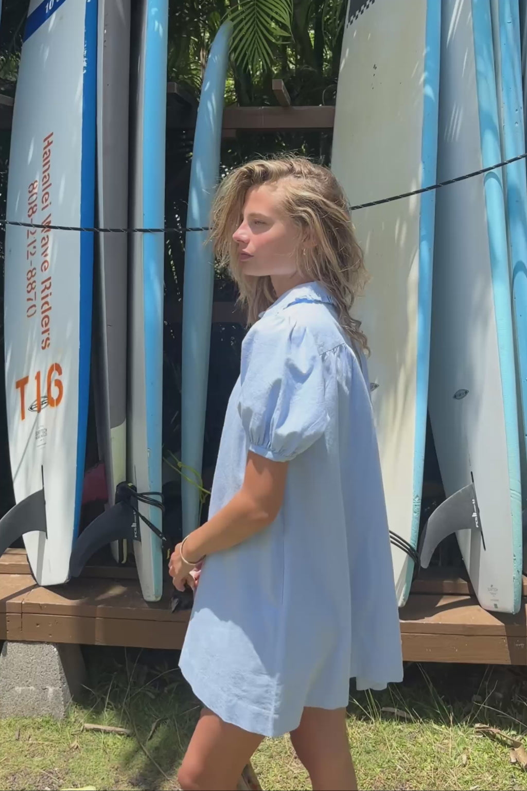 A video showing a woman in a light blue button-down chambray dress standing in front of kayaks with a woven bag in her hand