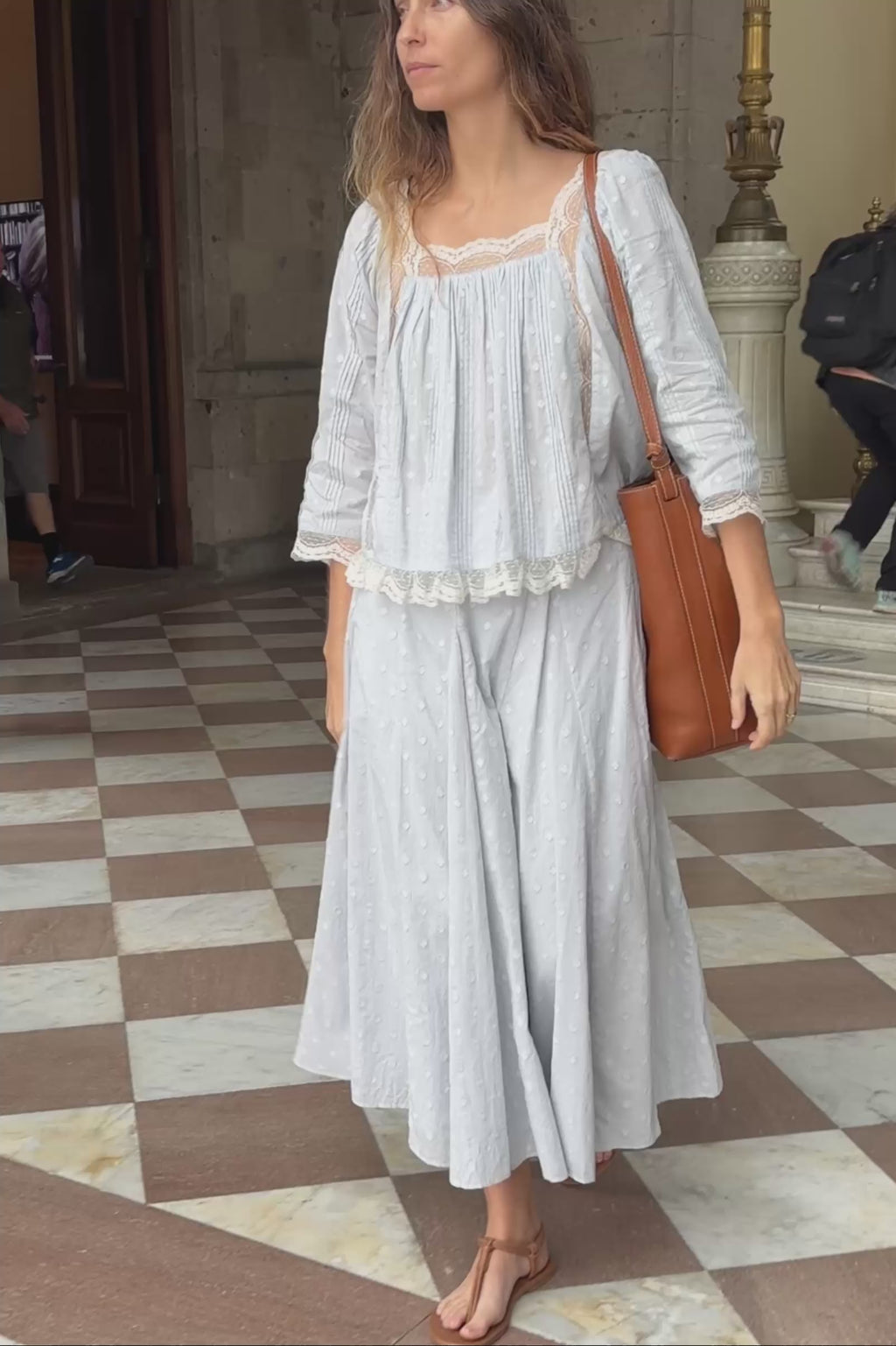 Woman in a light blue outfit walking in an elegant interior setting with a checkered floor.