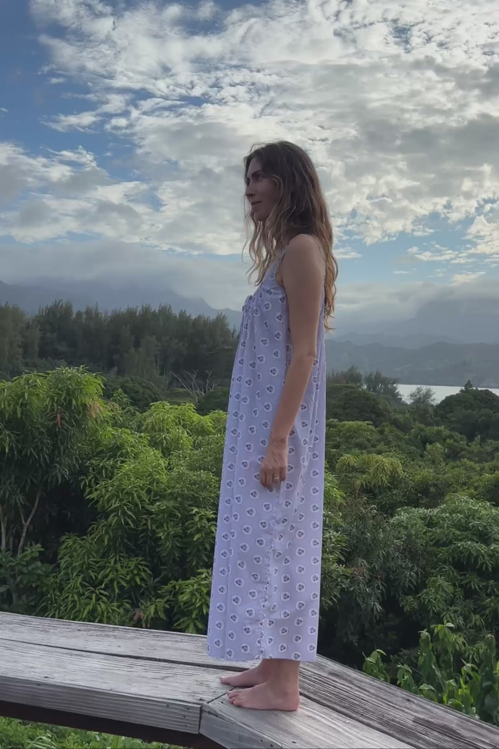 Woman in a light blue floral dress standing on a bench with greenery in the background