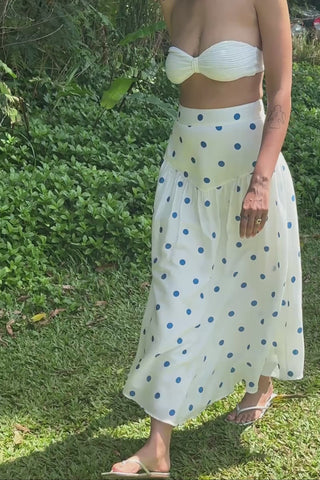 Woman in a white polka dot dress standing in a lush green garden with palm trees.
