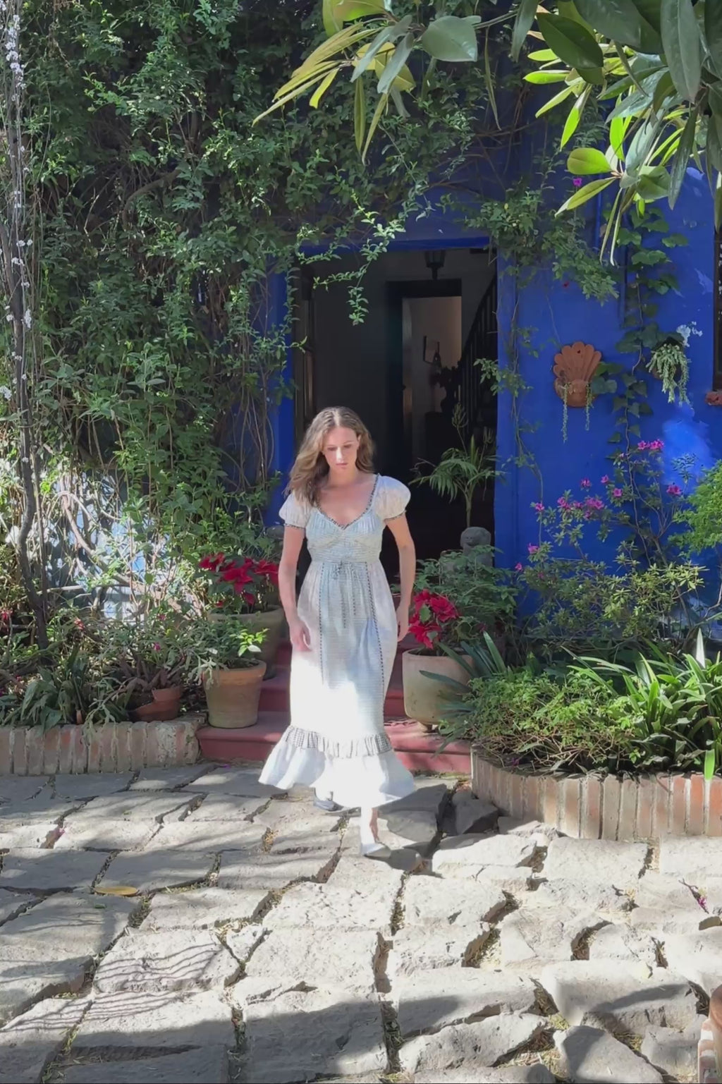 Woman in a patterned dress standing in a garden with potted plants and a colorful mural.