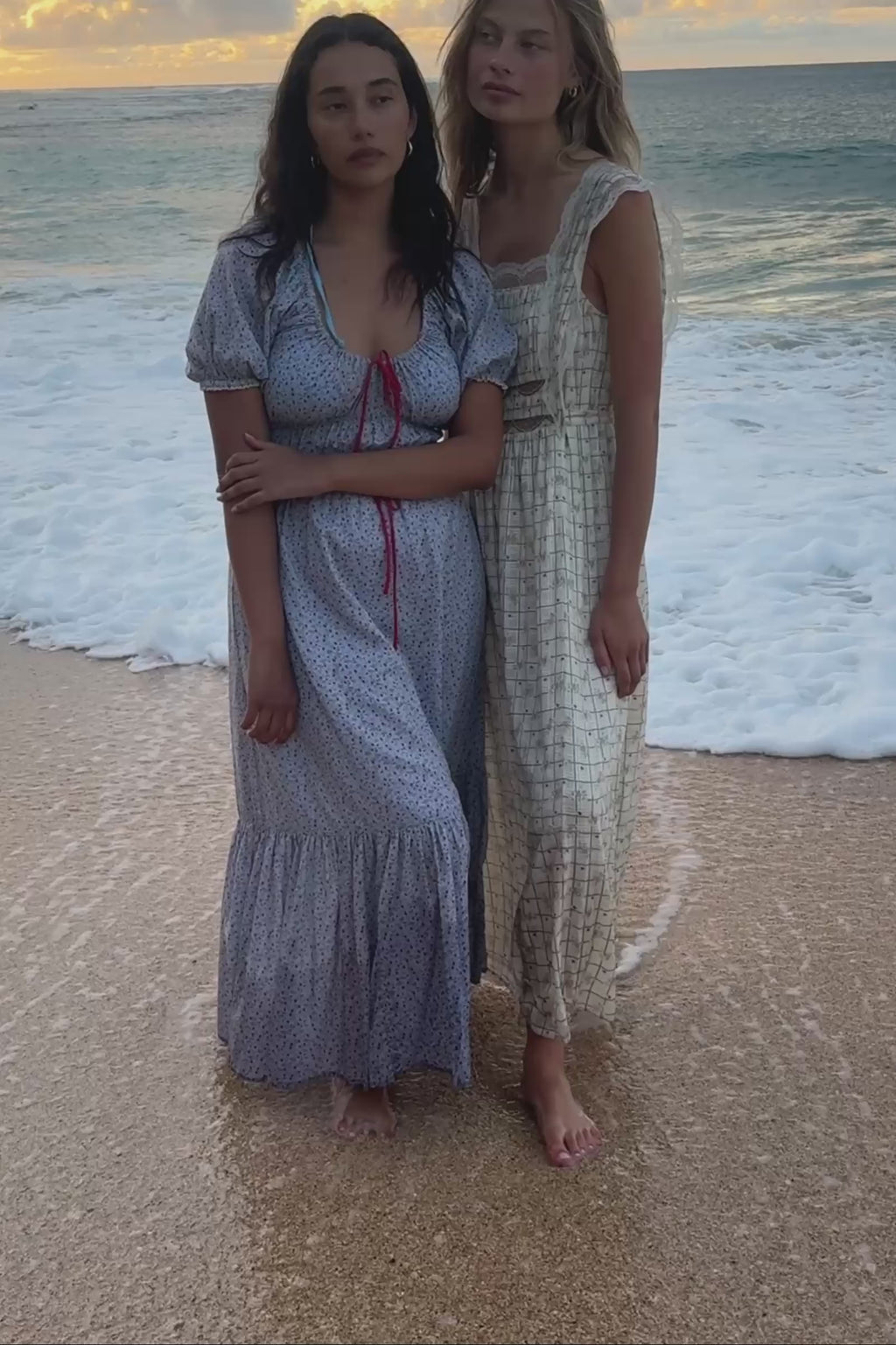 A video of two women standing in the waves, one in a light blue floral dress and another in a white patterned dress with lace.
