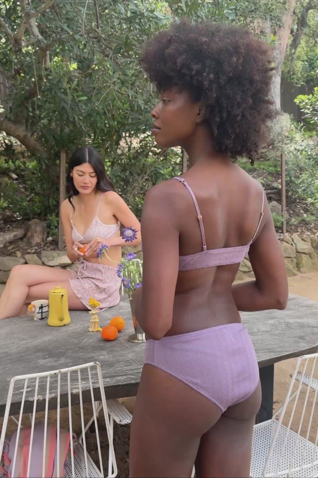 Woman in purple lingerie standing by a table with fruit and flowers outdoors.